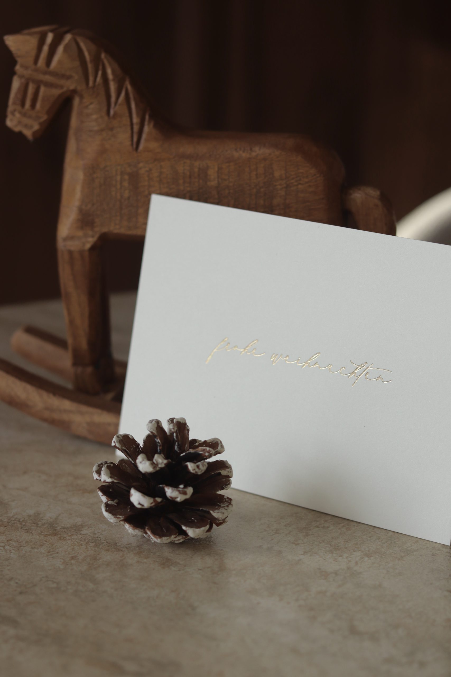 Weihnachtskarte im Querformat mit goldener Heißfolienprägung „Frohe Weihnachten“, dekoriert mit einem kleinen Holzschaukelpferd im Hintergrund.