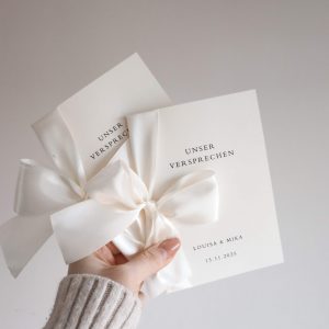 zwei Ehegelübdehefte mit weißer Schleife von einer Hand gehalten vor neutralem Hintergrund