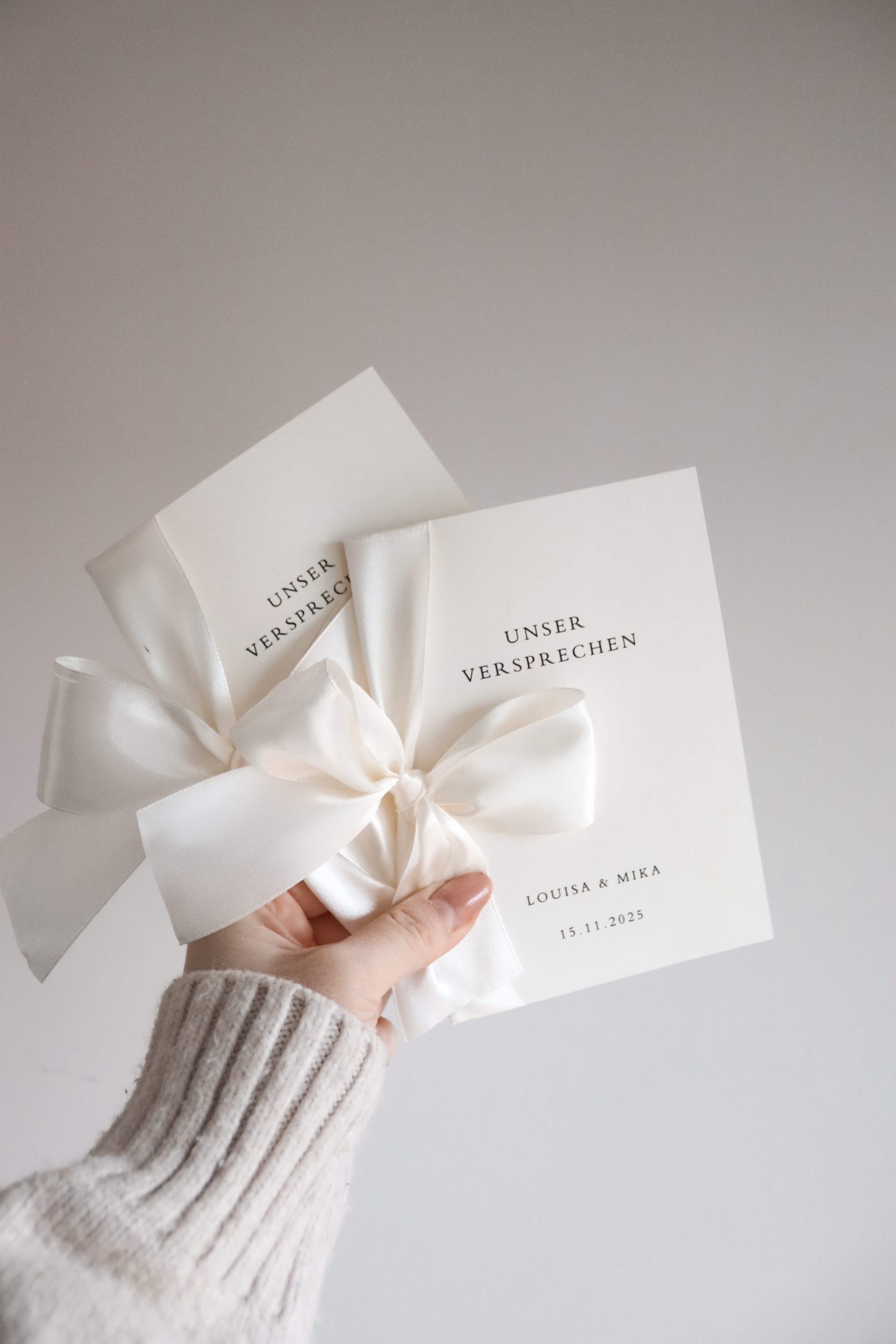 zwei Ehegelübdehefte mit weißer Schleife von einer Hand gehalten vor neutralem Hintergrund