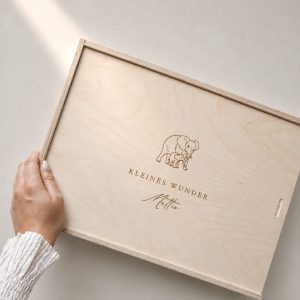 Minimalistische Aufnahme einer hellen Holzbox mit Gravur ‚Matteo‘ und Elefanten-Motiv auf einem hellen Greige-Hintergrund mit einem sanften Sonnenstrahl.