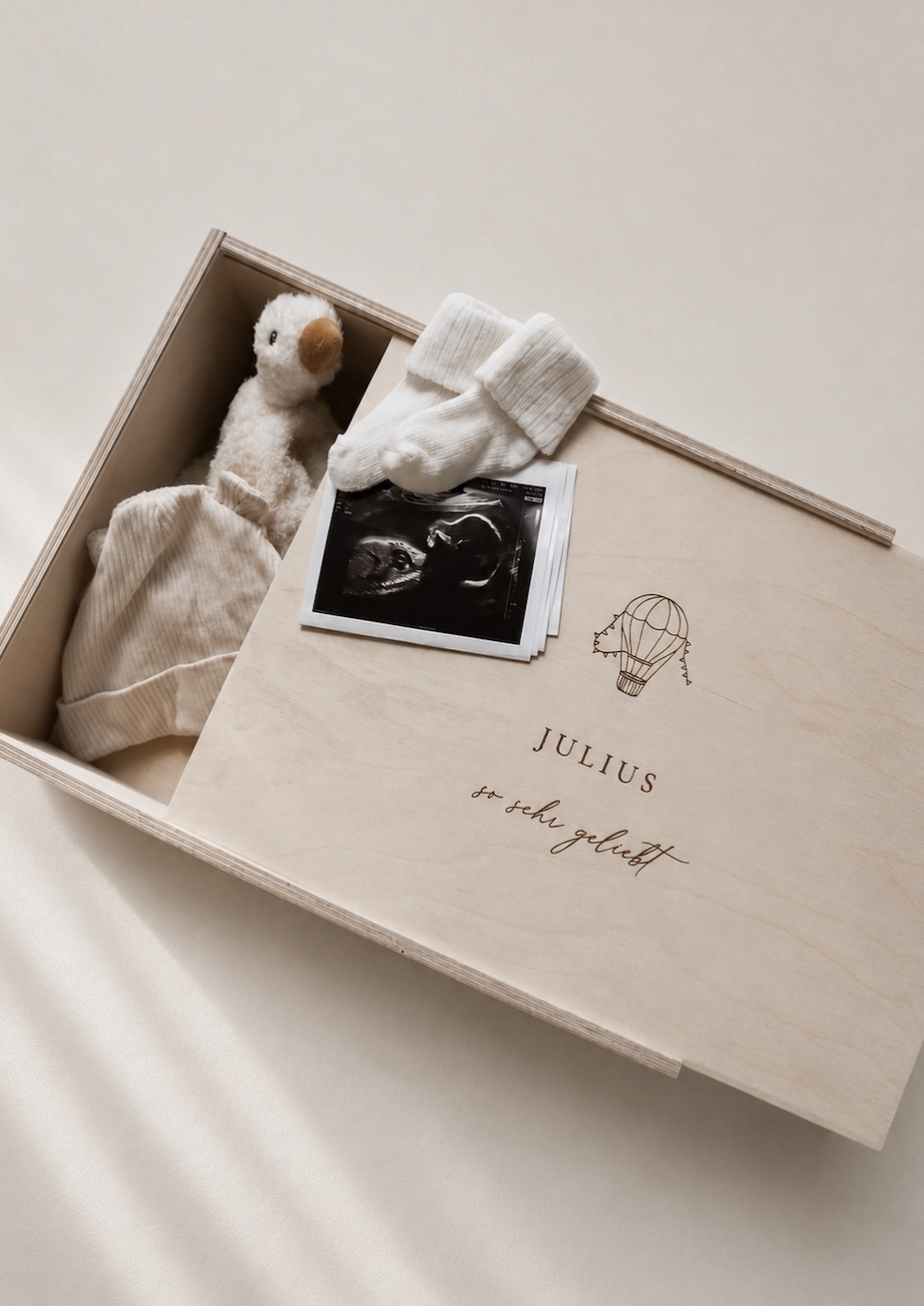 Minimalistische Aufnahme einer hellen Holzbox mit Gravur ‚JULIUS‘ und Heißluftballon-Motiv auf einem hellen Greige-Hintergrund mit einem sanften Sonnenstrahl.
