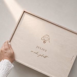 Minimalistische Aufnahme einer hellen Holzbox mit Gravur ‚JULIUS‘ und Heißluftballon-Motiv von einer Hand gehalten auf einem hellen Greige-Hintergrund mit einem sanften Sonnenstrahl.