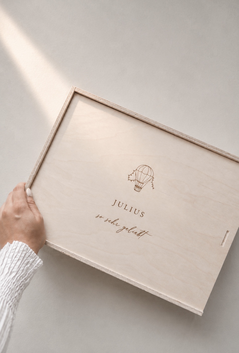 Minimalistische Aufnahme einer hellen Holzbox mit Gravur ‚JULIUS‘ und Heißluftballon-Motiv von einer Hand gehalten auf einem hellen Greige-Hintergrund mit einem sanften Sonnenstrahl.