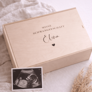 Erinnerungskiste aus hellem Buchenholz mit Schiebedeckel und gravierten Namen und Text "Meine SCHWANGERSCHAFT" auf einer hellen Decke und einem Ultraschallbild
