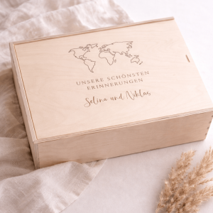 Erinnerungskiste aus hellem Buchenholz mit Schiebedeckel und gravierten Namen und Weltkarte auf einer hellen Decke