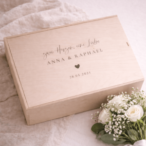 Erinnerungskiste aus hellem Buchenholz mit Schiebedeckel und gravierten Namen und Text "Zwei Herzen eine Liebe. Anna und Raphael. 28.05.2021" auf einer hellen Decke und einem Blumenstrauß