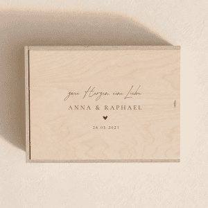 Personalisierte Erinnerungsbox aus hellem Holz zur Hochzeit mit eingraviertem Text "Zwei Herzen, eine Liebe" und Namen auf einem minimalistischen, cremefarbenen Hintergrund mit weichem Tageslicht.