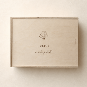 Personalisierte Erinnerungsbox aus hellem Holz mit eingraviertem Heißluftballonmotiv und Namen auf einem minimalistischen, cremefarbenen Hintergrund mit weichem Tageslicht.