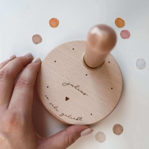 Runder Holzteller mit brauner Kerze und der Gravur "Julius - so sehr geliebt" umgeben von buntem Konfetti