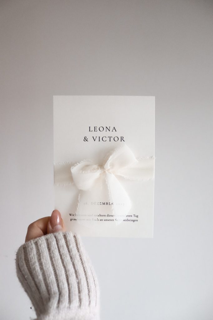 Eine Karte mit Druchbuchstabentext und weißer Schleife von einer Hand gehalten vor einem neutralen Hintergrund