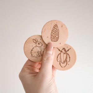 drei runde Holzscheiben mit den Motiven Hase, Tannenzapfen und Käfer von einer Hand gehalten vor einem neutralen Hintergrund