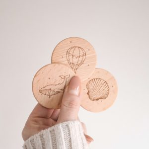 drei runde Holzscheiben mit den Motiven Wal, Muschel und Heißluftballon von einer Hand gehalten vor einem neutralen Hintergrund