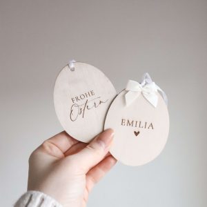 zwei Holzdekorationen in Form eines Ostereis mit dem Text "Emilia" und ein Herz und "Frohe Ostern", eine weiße Schleife oben, von einer Hand gehalten vor neutralem Hintergrund