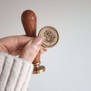 goldenes Wachssiegel mit dem Motiv eines Olivenzweigs und goldener Messingkopf an einem hölzernene Griff, gehalten von einer Hand vor einem neutralen Hintergrund