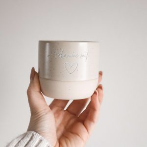 Beige runde Tasse ohne Griff aus Keramik und mit der Gravur "Hebamme mit" und dem Symbol "Herz" von einer Hand haltend vor einem neutralen Hintergrund