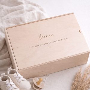 Erinnerungskiste aus hellem Buchenholz mit Schiebedeckel und gravierten Namen und Geburtsdaten auf einer hellen Decke und Babyschuhen und einem Schnuller