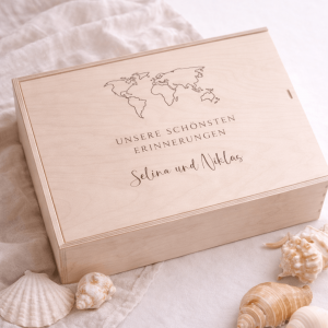 Erinnerungskiste aus hellem Buchenholz mit Schiebedeckel und gravierten Namen und Weltkarte auf einer hellen Decke und Muscheln zur Dekoration