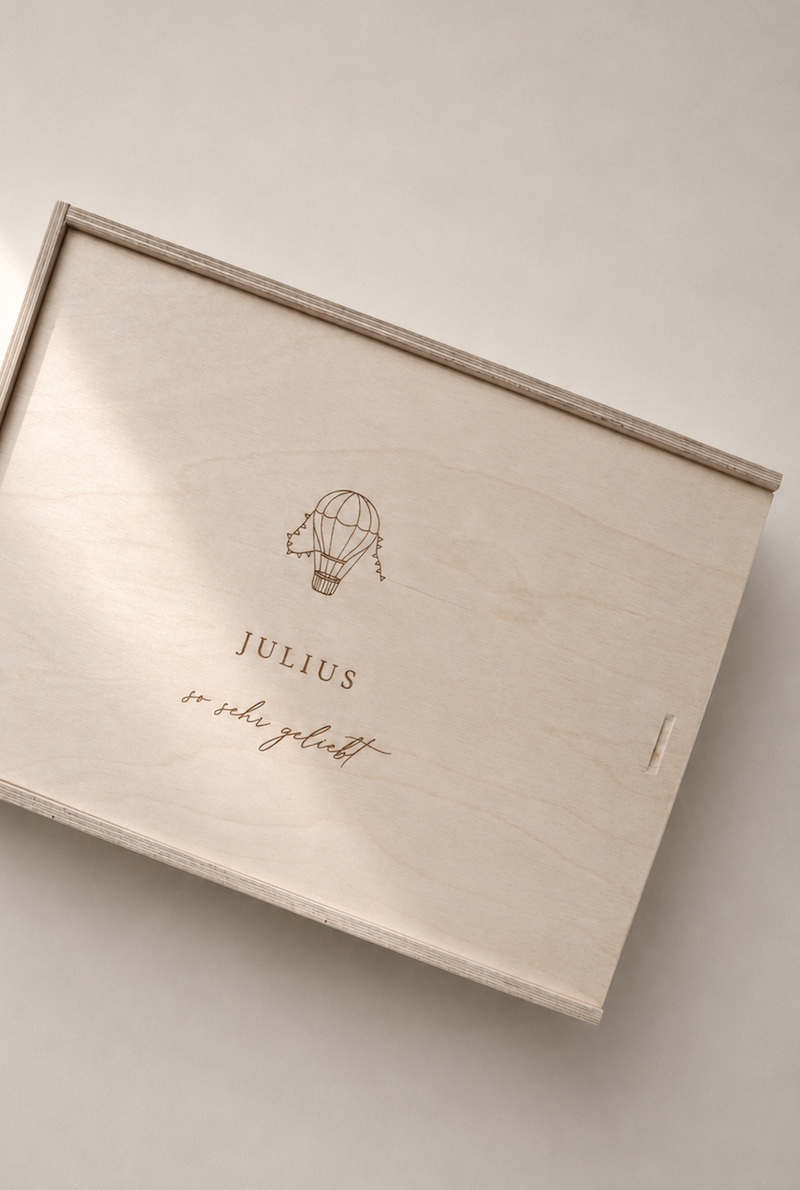 Minimalistische Aufnahme einer hellen Holzbox mit Gravur ‚JULIUS‘ und Heißluftballon-Motiv auf einem hellen Greige-Hintergrund mit einem sanften Sonnenstrahl.