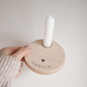 Runder Teller aus hellem Buchenholz mit der Gravur "Glückslicht" mit einem Herz darüber und einer weißen Kerze, von einer Hand gehalten vor einem neutralen Hintergrund