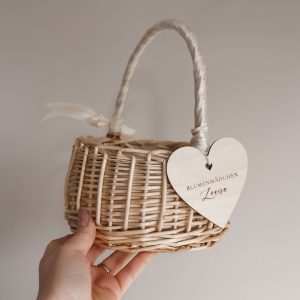 Personalisiertes Körbchen für Blumenmädchen aus geflochtenem Holz mit Henkel und Herzanhänger aus Holz mit graviertem Namen, in der Hand gehalten vor neutralem Hintergrund.