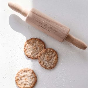 Kleines Wellholz aus Holz mit Gravur "minibäckerin marlene" mit drei hellen Keksen mit Puderzucker in minimalistischem Hintergrund mit natürlichem Sonnenlicht