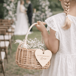 Mädchen in weißem Kleid mit einem Blumenkörbchen mit weißen Blumen und Herzanhänger mit Namen in einer Hochzeitsszenerie
