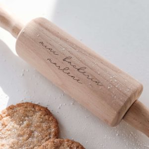 Kleines Wellholz aus Holz mit Gravur "minibäckerin marlene" mit drei hellen Keksen mit Puderzucker in minimalistischem Hintergrund mit natürlichem Sonnenlicht