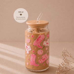 Glas mit Bambusdeckel und Strohhalm, gefüllt mit Eiskaffee, verziert mit sommerlichem pinken Cowgirl-Design und weißen Blüten, auf hellem Holztisch in warmem, minimalistischem Setting