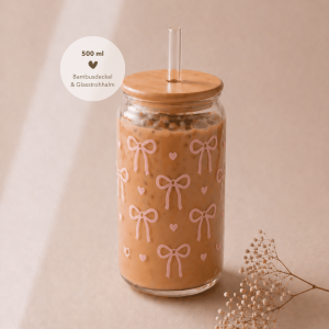 Glas mit Bambusdeckel und Strohhalm, gefüllt mit Eiskaffee, verziert mit sommerlichem rosa Schleifen-Design und rosa Herzen, auf hellem Holztisch in warmem, minimalistischem Setting