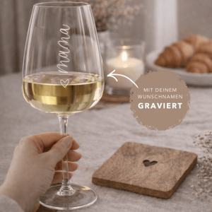 Personalisiertes Weinglas mit graviertem Namen „Mama“, gefüllt mit Weißwein, in der Hand gehalten vor dekorativem Tisch mit Kerze und Croissants im Hintergrund