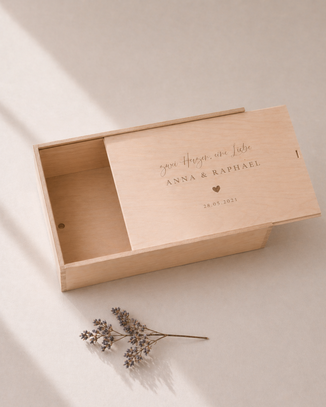 Minimalistische Aufnahme einer personalisierten Holz-Erinnerungskiste mit Gravur „Zwei Herzen eine Liebe, Anna und Raphael“ und Datum, auf hellem Hintergrund mit sanftem Sonnenlicht und einem kleinen getrockneten Zweig im Vordergrund.