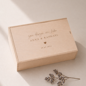 Minimalistische Aufnahme einer personalisierten Holz-Erinnerungskiste mit Gravur „Zwei Herzen eine Liebe, Anna und Raphael“ und Datum, auf hellem Hintergrund mit sanftem Sonnenlicht und einem kleinen getrockneten Zweig im Vordergrund.