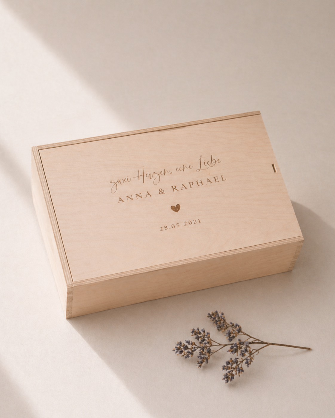 Minimalistische Aufnahme einer personalisierten Holz-Erinnerungskiste mit Gravur „Zwei Herzen eine Liebe, Anna und Raphael“ und Datum, auf hellem Hintergrund mit sanftem Sonnenlicht und einem kleinen getrockneten Zweig im Vordergrund.
