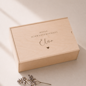 Minimalistische Aufnahme einer personalisierten Holz-Erinnerungskiste mit Gravur „meine schwangerschaft Elena" und einem Herz, auf hellem Hintergrund mit sanftem Sonnenlicht und einem kleinen getrockneten Zweig im Vordergrund.