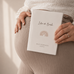 Eine schwangere Frau mit Hand auf dem Bauch und einer personalisierten Mutterpasshülle mit dem Schriftzug "Liebe im Bauch" und einem Regenbogen