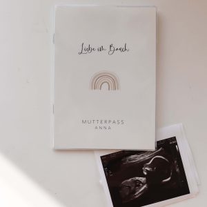 Weiße Mutterpasshülle mit dem Text "Liebe im Bauch" und "Mutterpass Anna" und der Aquarellzeichnung eines Regenbogens, daneben ein Ultraschallbild in minimalistischem Hintergrund