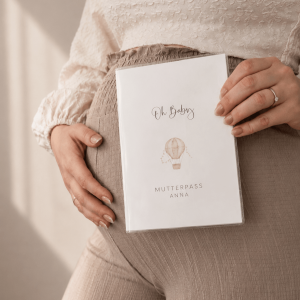 Eine schwangere Frau mit Hand auf dem Bauch und einer personalisierten Mutterpasshülle mit dem Schriftzug "Oh Baby" und einem Heißluftballon