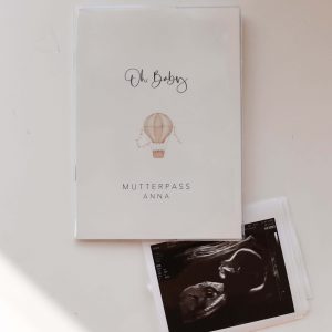 Weiße Mutterpasshülle mit dem Text "Oh Baby" und "Mutterpass Anna" und der Aquarellzeichnung eines Heißluftballons, daneben ein Ultraschallbild in minimalistischem Hintergrund