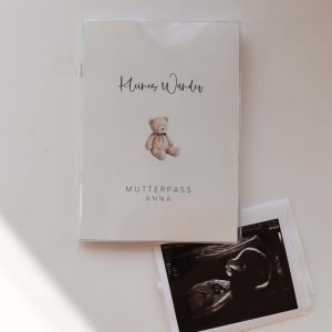 Weiße Mutterpasshülle mit dem Text "Kleines Wunder" und "Mutterpass Anna" und der Aquarellzeichnung eines Teddybären, daneben ein Ultraschallbild in minimalistischem Hintergrund