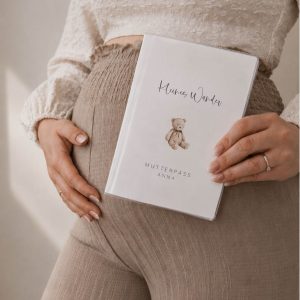 Eine schwangere Frau mit Hand auf dem Bauch und einer personalisierten Mutterpasshülle mit dem Schriftzug "Kleines Wunder" und einem Teddybären