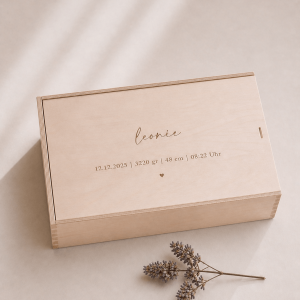 Minimalistische Aufnahme einer personalisierten Holz-Erinnerungskiste mit Gravur „Leonie“ und Datum, auf hellem Hintergrund mit sanftem Sonnenlicht und einem kleinen getrockneten Zweig im Vordergrund.