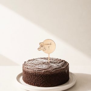 Großer Cake Topper aus Lindenholz mit Bären-Motiv, dem Namen noah und der Zahl 1 in einem Schokokuchen in minimalistischem Hintergrund mit natürlichem Sonnenlicht
