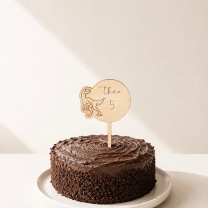 Großer Cake Topper aus Lindenholz mit Dinosaurier-Motiv, dem Namen Theo und der Zahl 5 in einem Schokokuchen in minimalistischem Hintergrund mit natürlichem Sonnenlicht