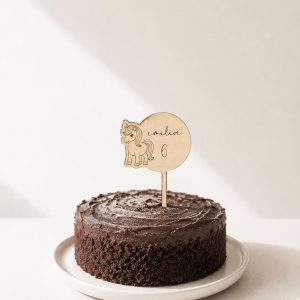 Großer Cake Topper aus Lindenholz mit Einhorn-Motiv, dem Namen Emilia und der Zahl 6 in einem Schokokuchen in minimalistischem Hintergrund mit natürlichem Sonnenlicht