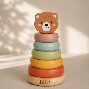Personalisierter Holzsteckturm mit fünf bunten Stapelringen und niedlichem Bärenkopf aus Holz. Der Name „JULIUS“ ist auf dem Holzsockel eingraviert. Das Motorikspielzeug steht vor einem hellen Hintergrund mit weichen Licht- und Schatteneffekten.