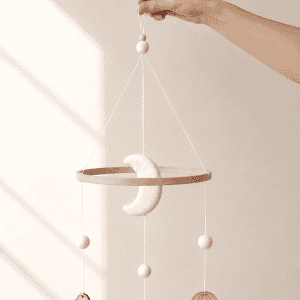 Handgefertigtes Babymobile aus Holz und Filz mit Mond, Wolke, Vogel und Heißluftballon. Das Mobile wird an einer kleinen Schlaufe gehalten und vor einem minimalistischen Hintergrund mit natürlichem Sonnenlicht präsentiert – ideal als zeitlose Dekoration für das Babyzimmer.