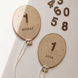 zwei Luftballons aus Holz mit weißer Kordel mit Zahlen und der Beschriftung "1 Monat" und "1 Jahr" auf natürlichem Untergrund