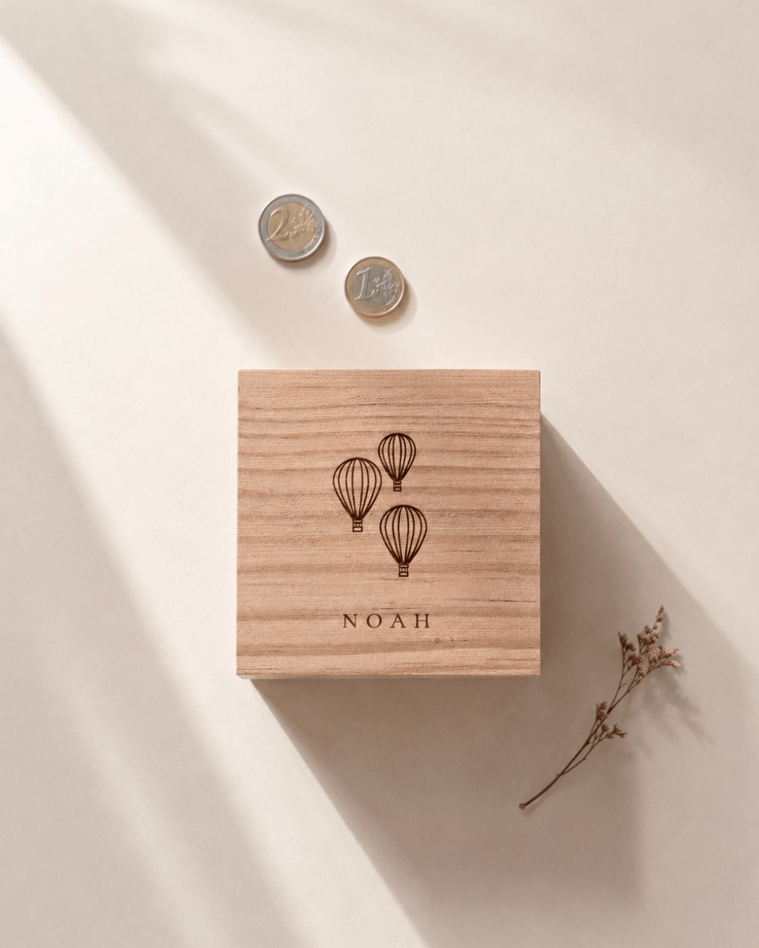 Quadratische Spardose aus Holz mit natürlicher Maserung und Motiv mit drei Heißluftballons und Namensgravur. In minimalistischem Hintergrund mit natürlichem Sonnenlicht