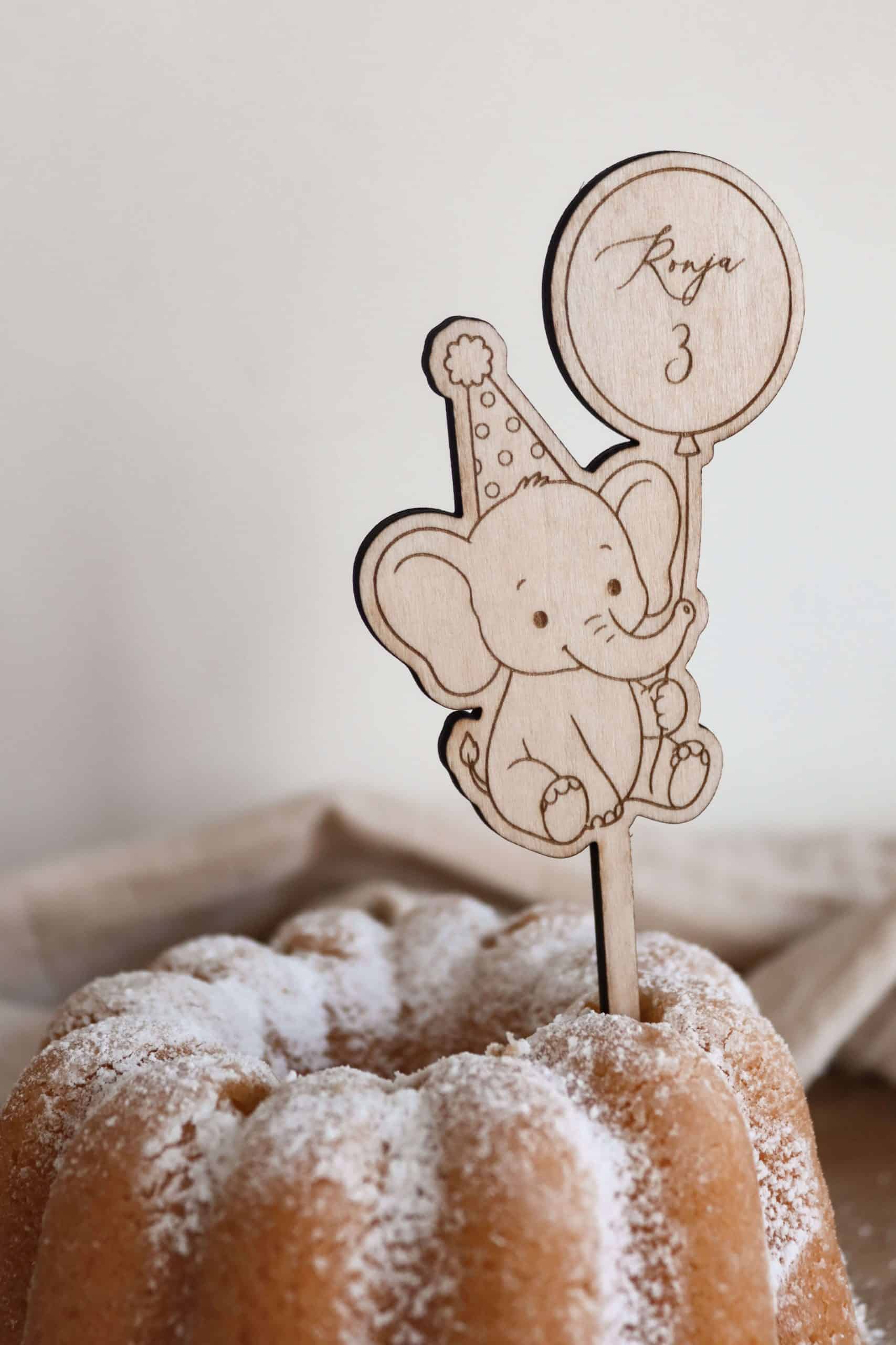 Das Bild zeigt einen Caketopper aus hellem Holz mit dem Motiv eines kindgerechten Elefanten mit Partyhut und einem Ballon in der Hand, welcher den Namen "Ronja" und die Zahl "3" ziert. Der Kuchenstecker steckt in einem hellen Kuchen Kuchen mit Puderzucker