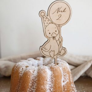 Das Bild zeigt einen Caketopper aus hellem Holz mit dem Motiv einer kindgerechten Gans mit Partyhut und einem Ballon in der Hand, welcher den Namen "Noah" und die Zahl "2" ziert. Der Kuchenstecker steckt in einem hellen Kuchen Kuchen mit Puderzucker